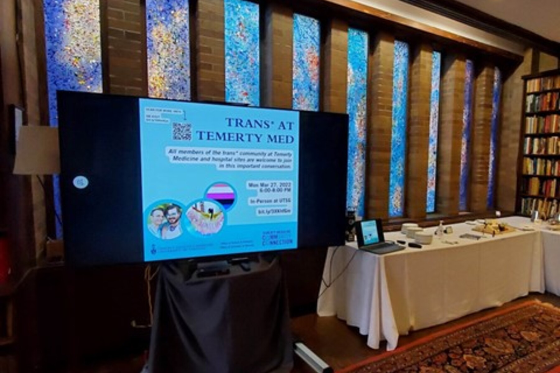 Photograph of a screen in a library showing a slide that reads: "Trans* at Temerty Med. All members of the trans* community of Temerty Medicine and hospital sites are welcome to join in this important conversation. Mon Mar 27, 2022 6:00 - 8:00PM In-person at UTSG" with a link to the event. 